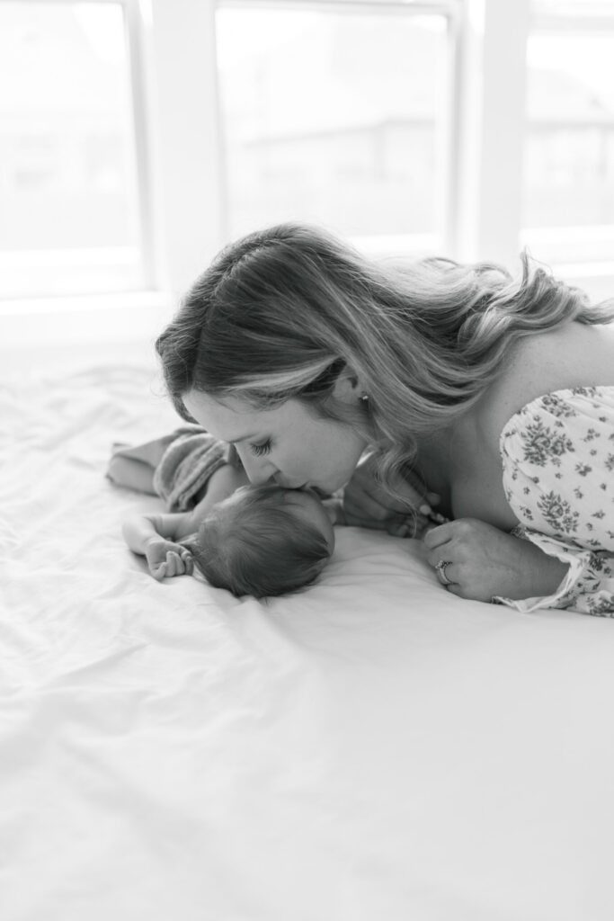 Mom leans over newborn baby and kisses his head as he sleeps on the bed.