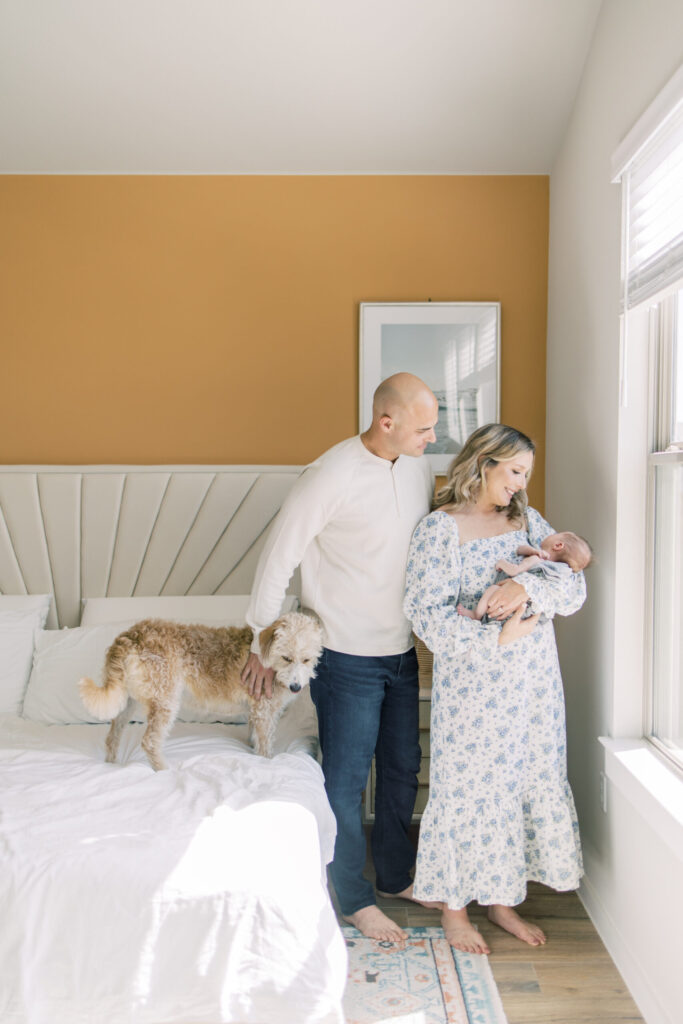 Parents admire their newborn in master bedroom with orange walls and dog on bed.