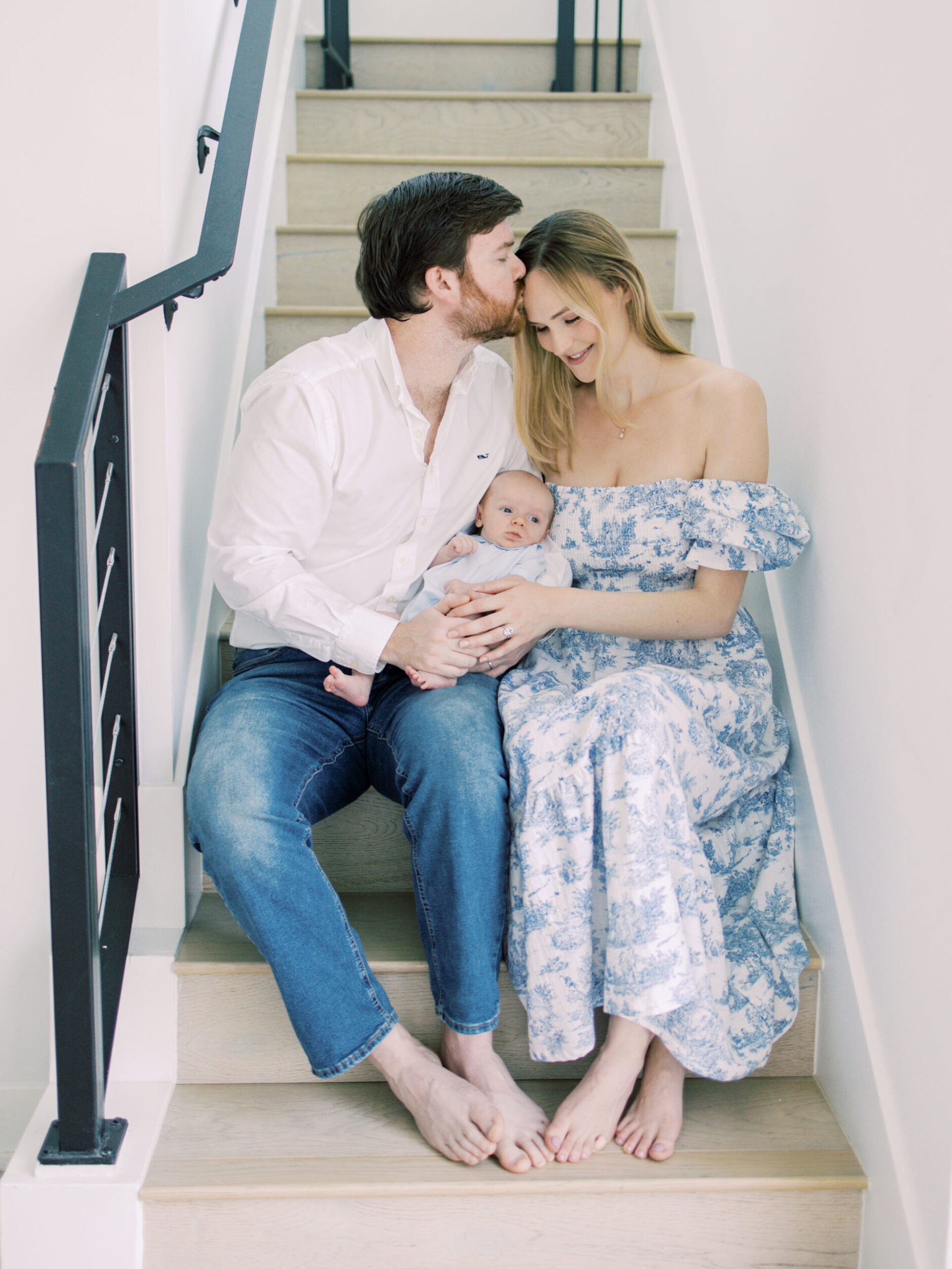 Husband gently kisses wife on head as they hold their newborn on the stairs.