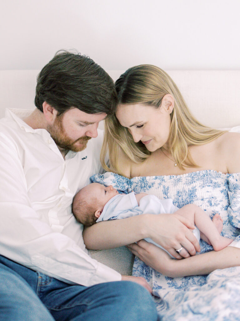 Couple smile adoringly at their newborn baby boy cradled in his mother's arms in newborn portraits.