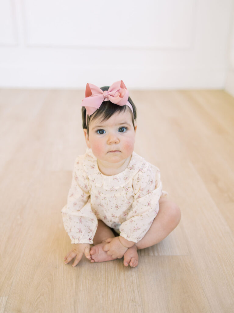 Baby girl sits up on hardwood floor in flower outfit.