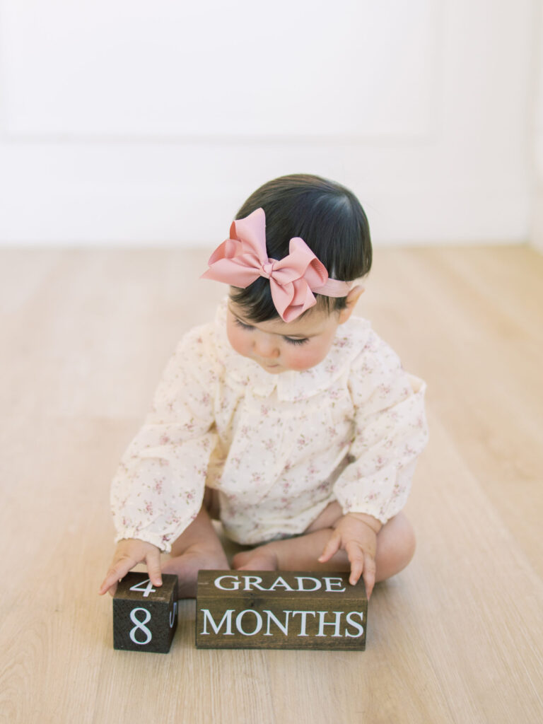 Baby girls sits up with and holds blocks that say 8 months.