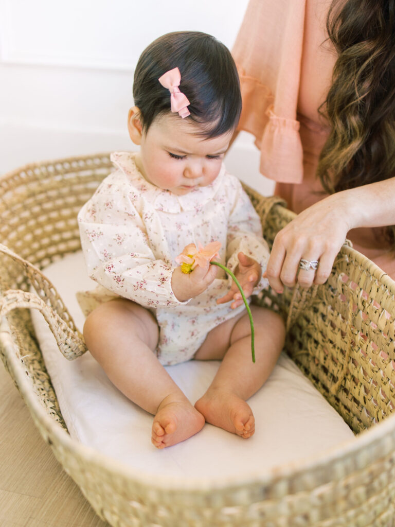 Baby girl holds flower in basket while sitting up.